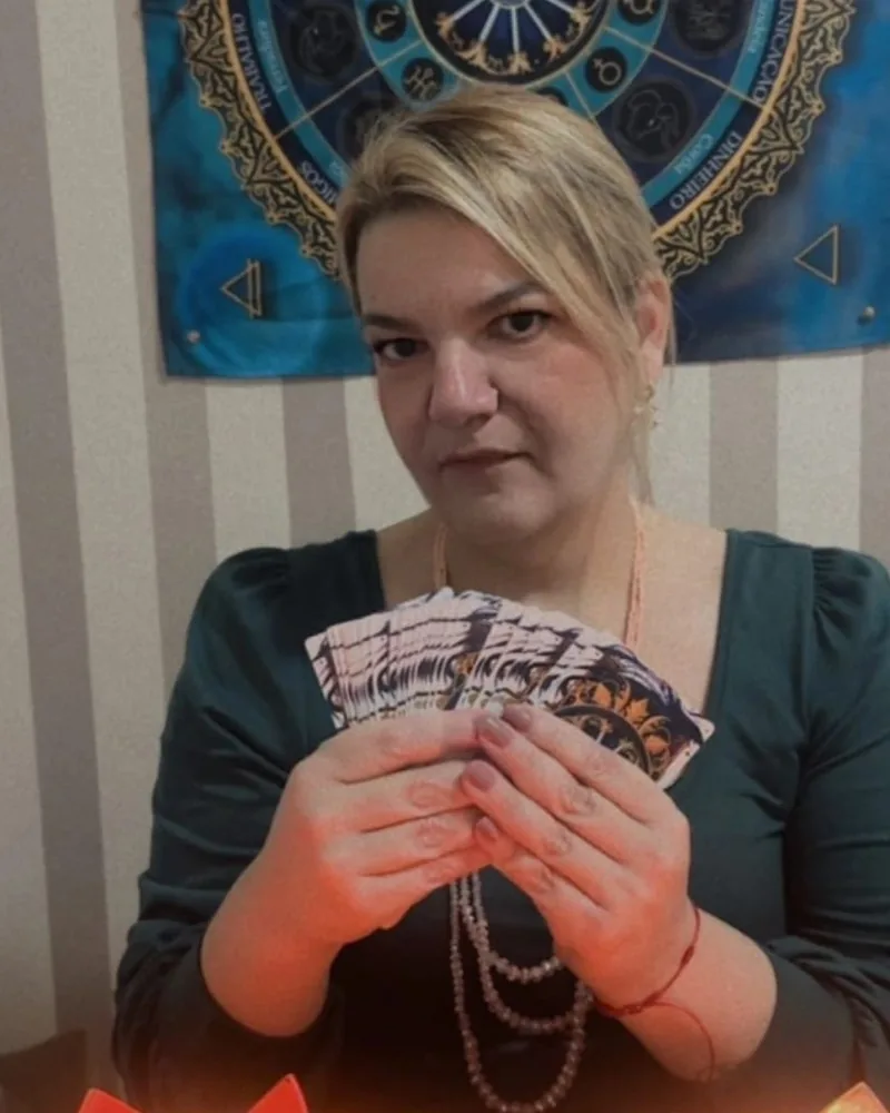 Cigana Laura segurando um leque de cartas do Baralho Cigano, com o painel de astrologia ao fundo